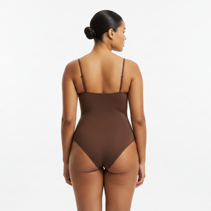 Professional 1:1 studio photo of the same woman. No change in face or body. Natural skin texture. Real pores. Natural imperfections. Thin shoulder straps. Shapewear bodysuit in bruin. Soft white studio lighting. Gentle shadow. Fashion photography look. Back view arms relaxed. Curvy. Warme bruine huid. Low bun. 26 jaar.