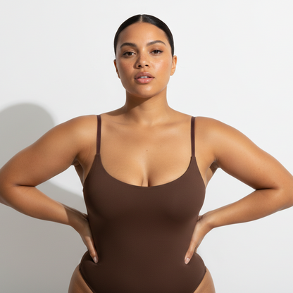 Professional 1:1 studiofoto van dezelfde vrouw. Geen verandering in gezicht of lichaam. Real skin texture. Real pores. Natural imperfections. Dunne schouderbandjes. Shapewear bodysuit in bruin. Clean white studio. Zachte schaduw. Direct eye contact. Pose handen in de zij met een lichte heupshift. Confident uitstraling. Curvy. Bruine huid. Low bun. 26 jaar.